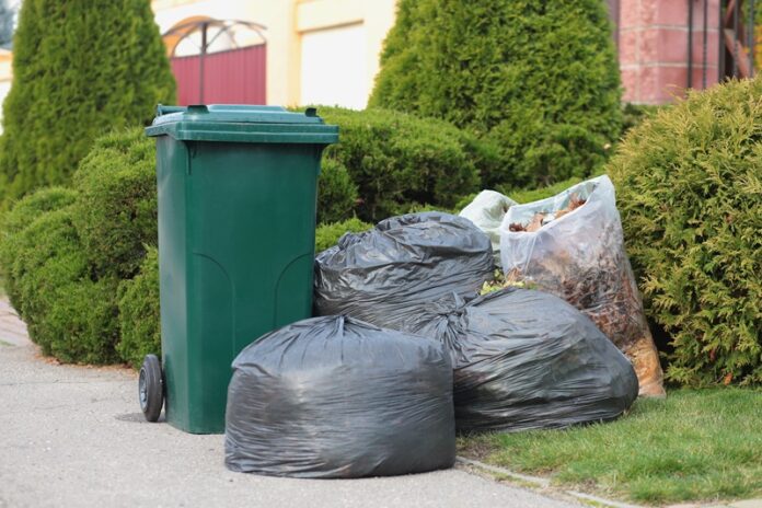 A large green plastic trash can in front of a modern house with green trees along with trash bags. Infection control, garbage disposal, waste recycling concept Automatyzacja, sztuczna inteligencja i bezpieczeństwo – rewolucja w branży gospodarowania odpadami